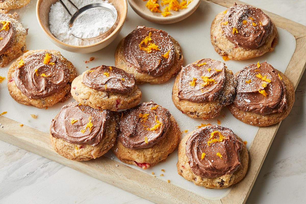 Overhead horizontal image of chocolate-covered cookies with a dusting of powdered sugar and orange zest on a parchment-lined wooden board. A bowl of powdered sugar with a sifter is also visible.