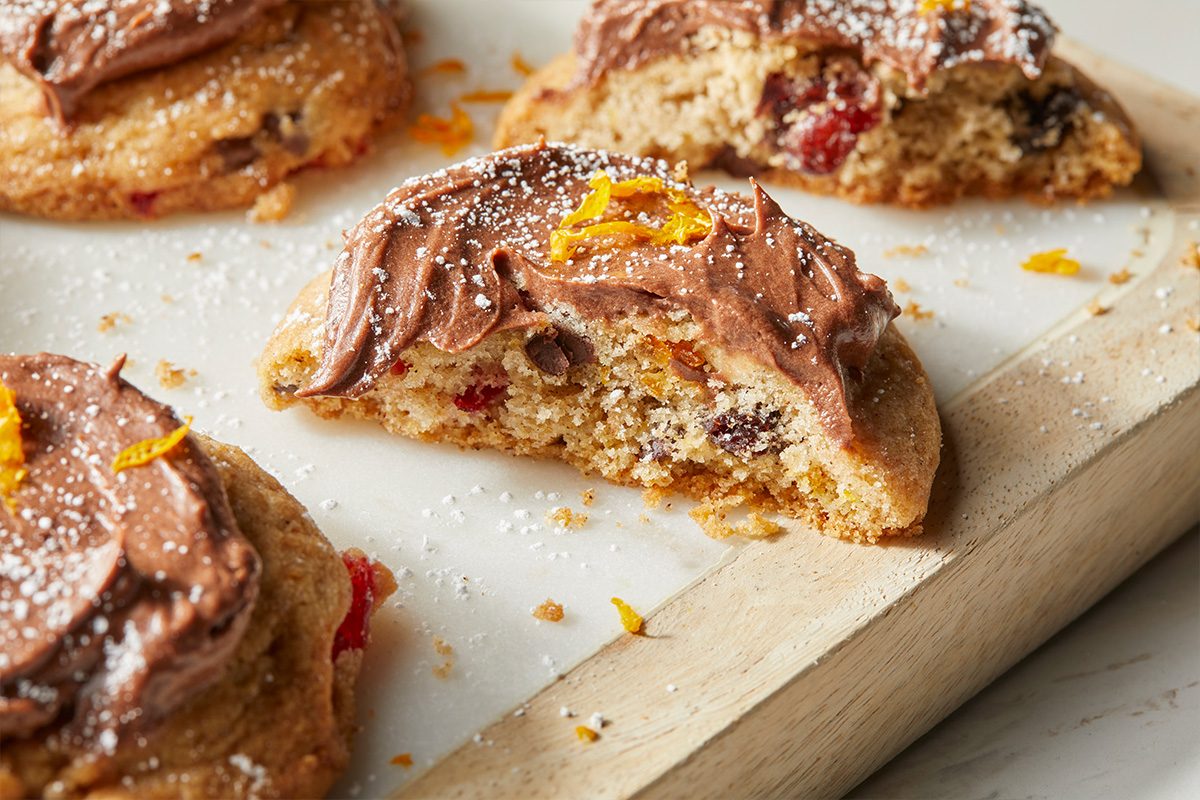 Eye Level horizontal image of baked cookies with frosting and powdered sugar, one of which has a bite taken out of it. The cookies contain chocolate chips, cherries and orange zest.
