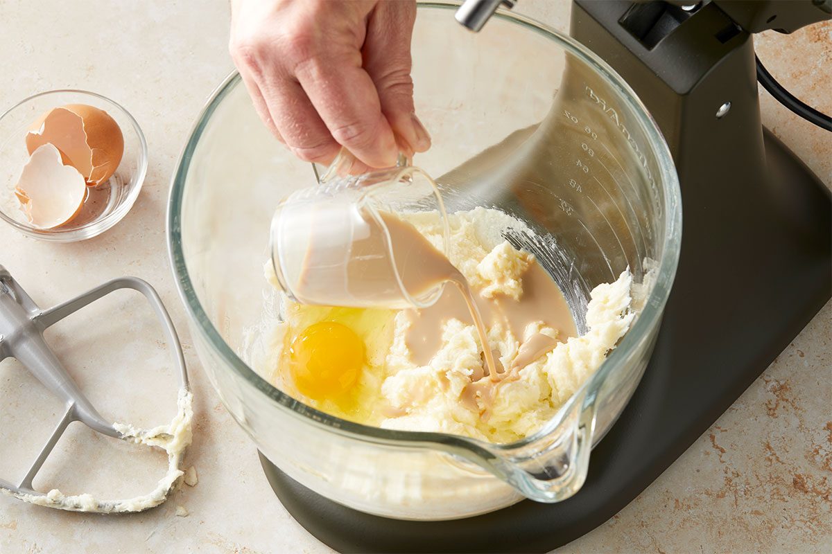 Eye Level horizontal image of ingredients being mixed in a glass bowl. A hand pours milk from a small glass container into a bowl that already contains butter and an egg. | [Original] Pouring Irish cream into bowl with beaten butter & sugar, egg already in bowl, not yet mixed in.