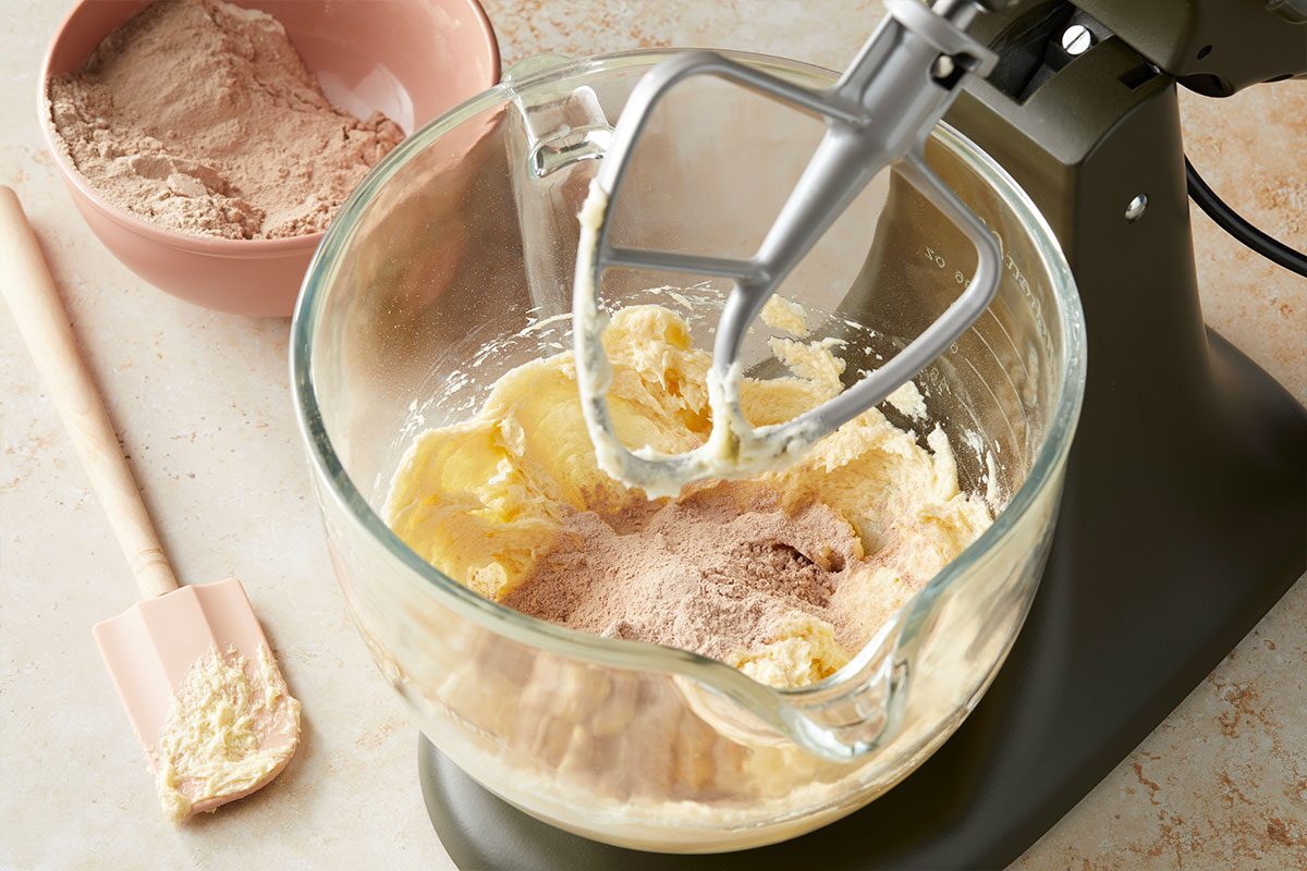 Eye Level horizontal image of ingredients being mixed in a clear glass bowl. A stand mixer with a beater attachment is mixing the ingredients. Additional powdered ingredients are sitting on top of the dough. | [Original] Adding dry ingredients to batter