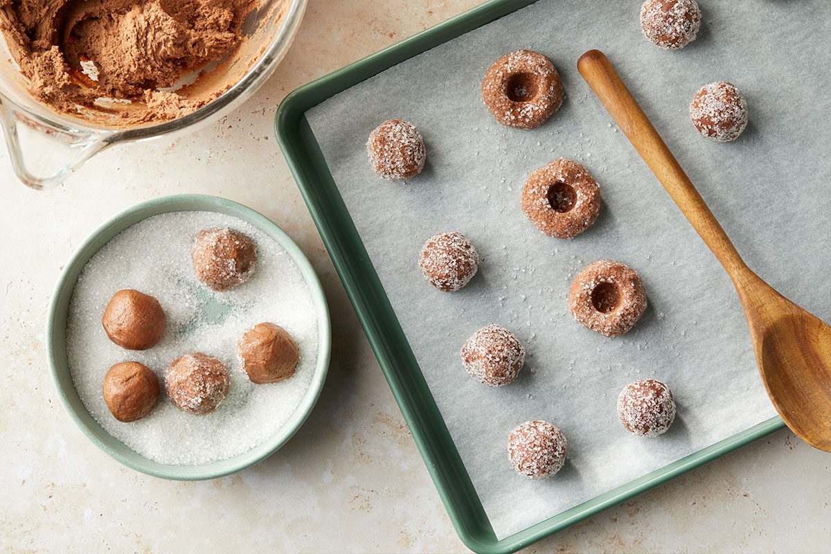 Overhead horizontal image of chocolate cookie dough being prepared. Some of the dough balls are coated in sugar, while others are uncoated, sitting in a bowl of sugar, and some are on a baking sheet, some with an indentation made with a wooden spoon. | [Original] Process shot – dough rolled into balls, some being rolled in sugar, some on parchment-lined baking sheet and indented with spoon