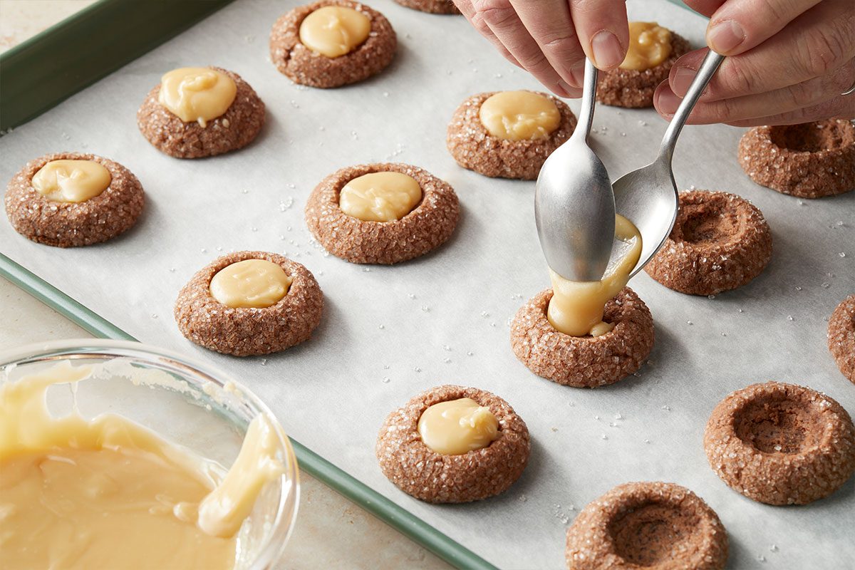 Overhead horizontal image of cookies being filled with a spoon. The cookies are on a baking sheet lined with parchment paper, and they are brown with sugar crystals around the edges. A bowl of filling is visible in the lower left corner. | [Original] Filling cookies with ganache