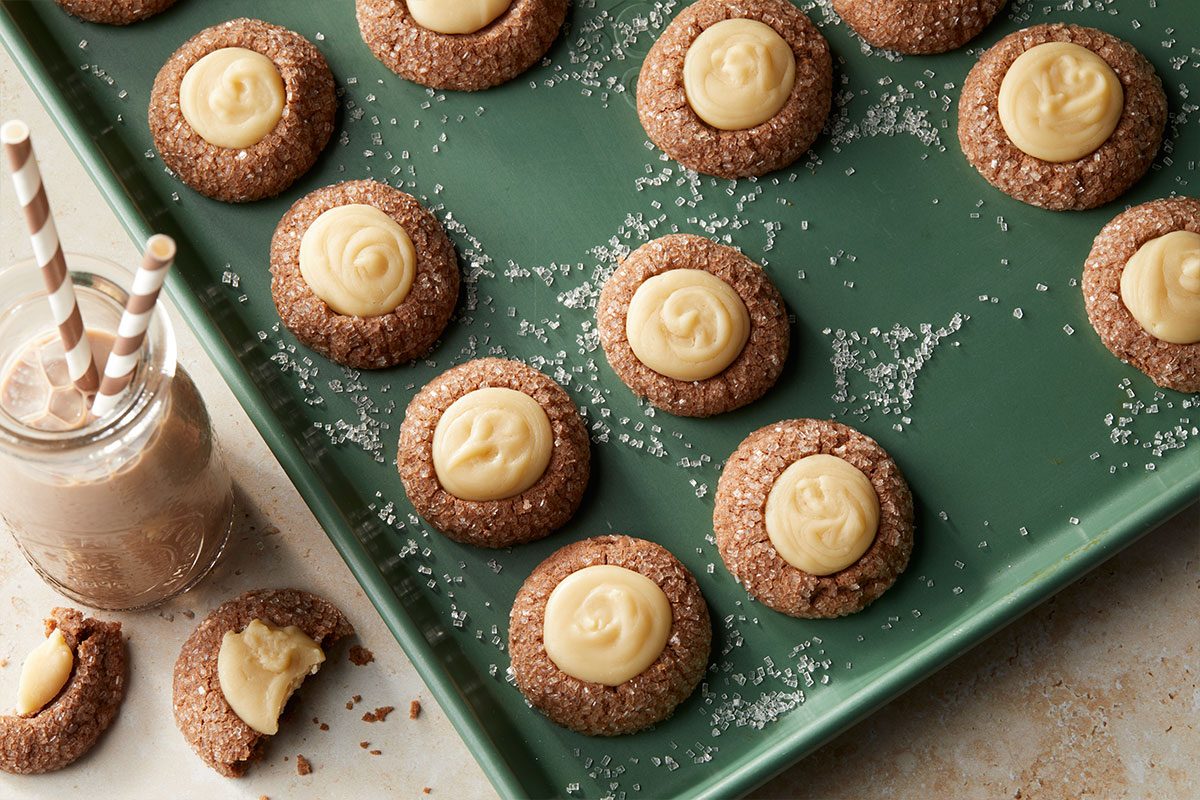Overhead horizontal image of thumbprint cookies on a green baking sheet, sprinkled with sugar. A glass of chocolate milk with two striped straws sits next to the baking sheet with one broken cookie.