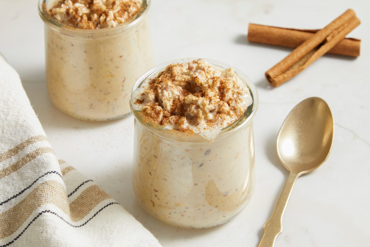 Eye Level horizontal image of overnight oats in a clear glass jar. The oats are topped with cinnamon and other jars, a spoon, cinnamon sticks, and a striped kitchen towel are also in the shot.