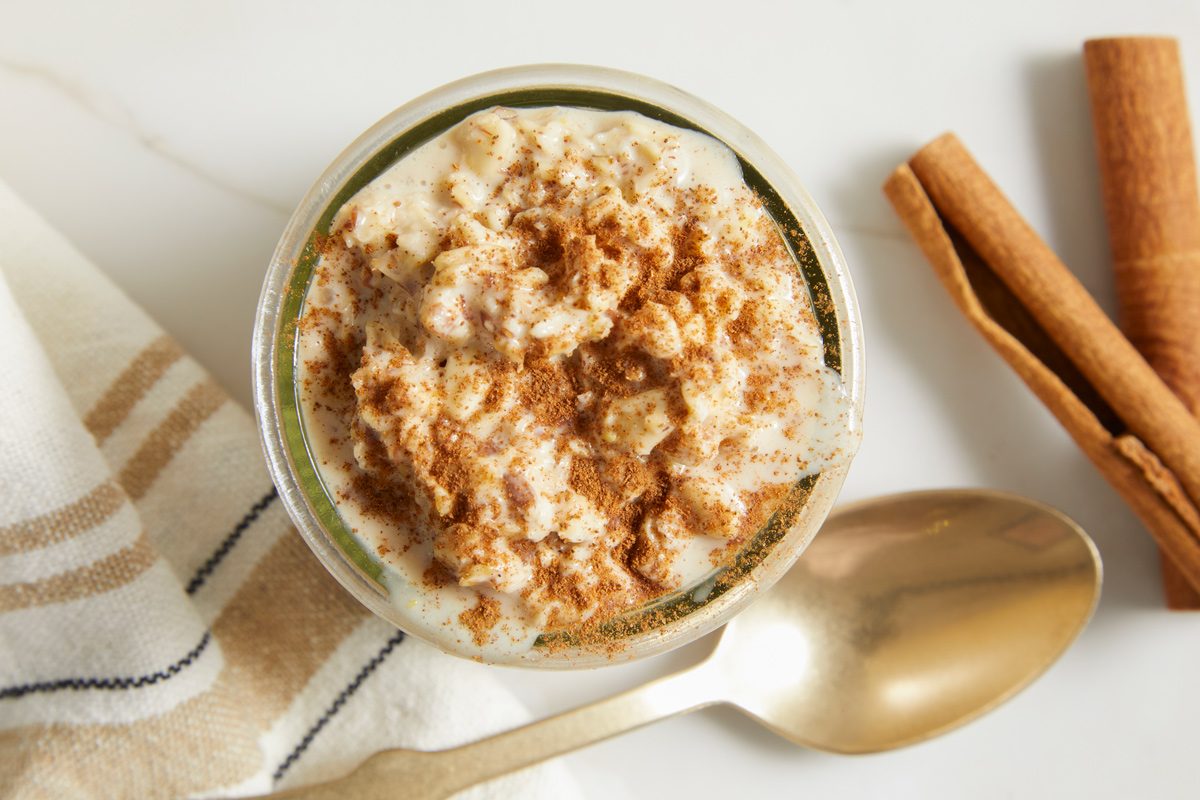 Overhead horizontal image of a glass jar filled with rice pudding sprinkled with cinnamon. A golden spoon rests beside the jar, and cinnamon sticks sit in the background.
