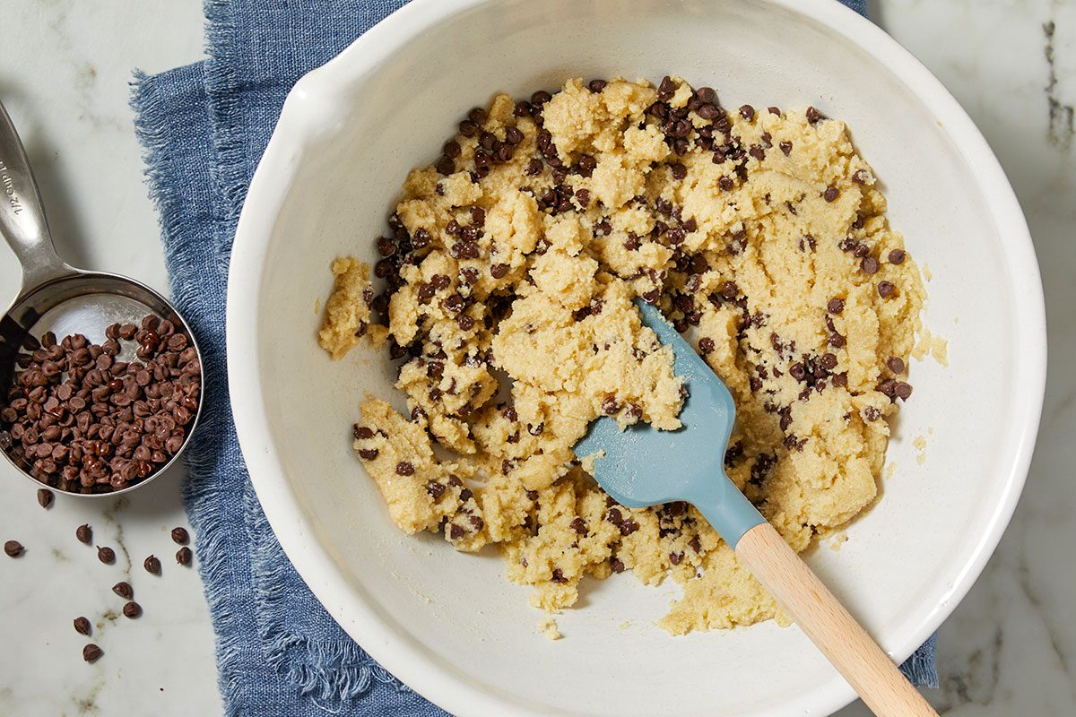 Overhead horizontal image of cookie dough in a white bowl with a blue spatula. Next to the bowl is a measuring cup full of chocolate chips on a blue linen. The cookie dough is clumpy and dotted with chocolate chips.