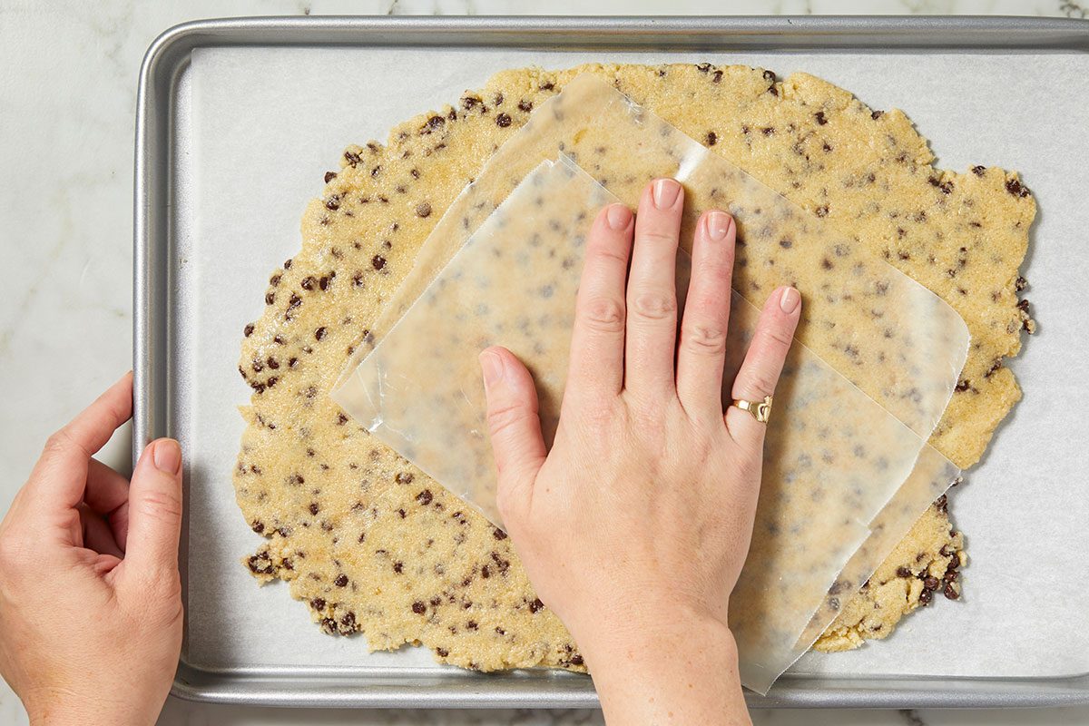 Overhead horizontal image of cookie dough being pressed out with parchment paper on a baking sheet. The dough has chocolate chips mixed in.