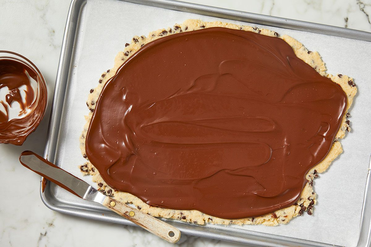 Overhead horizontal image of spreading melted chocolate over a cookie crust on a baking sheet. A bowl of melted chocolate and a spatula are visible next to the cookie.