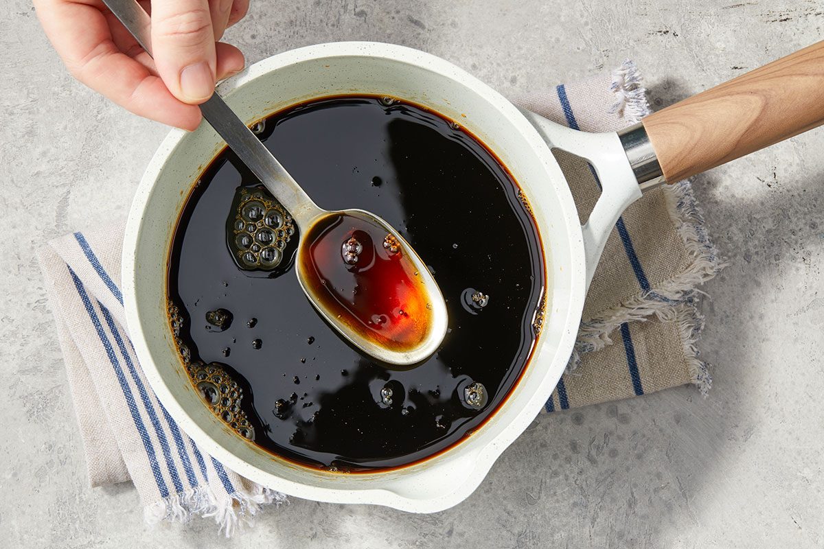 Overhead horizontal image of a hand stirring a dark sauce in a light-colored pan with a metal spoon. Bubbles are visible in the dark brown sauce.