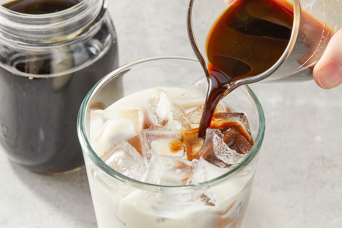 Eye Level horizontal image of someone pouring coffee into a glass. The glass is filled with milk and ice. A jar of coffee sits in the background.
