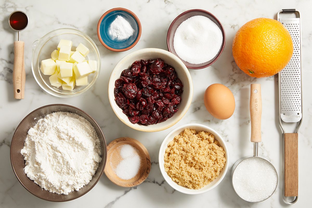 Baking ingredients arranged on a marble surface, including flour, brown sugar, butter, egg, dried cranberries, orange, grater, spices, baking powder, salt, and small bowls of sugar.