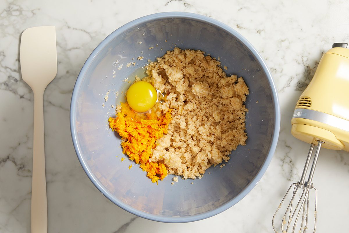 A mixing bowl containing cookie dough, an egg, and orange zest on a marble countertop. Next to the bowl are a spatula and a yellow electric hand mixer with beaters attached.