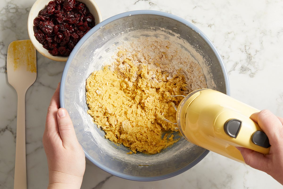 A person uses a hand mixer to blend yellow dough in a large bowl, with a bowl of dried cranberries and a spatula nearby on a marble countertop.