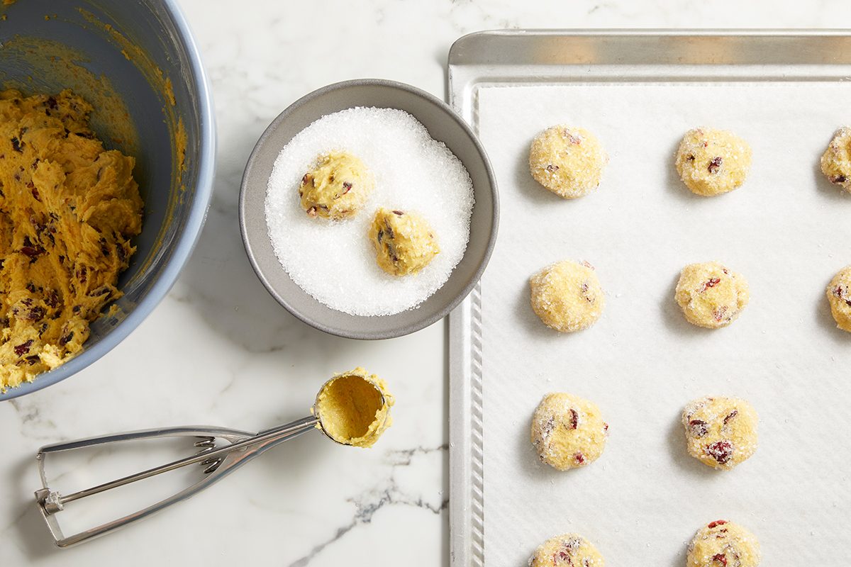 A baking sheet lined with parchment paper holds unbaked cookie dough balls. A mixing bowl with dough, a bowl of sugar, and a cookie scoop are also visible on a marble countertop.