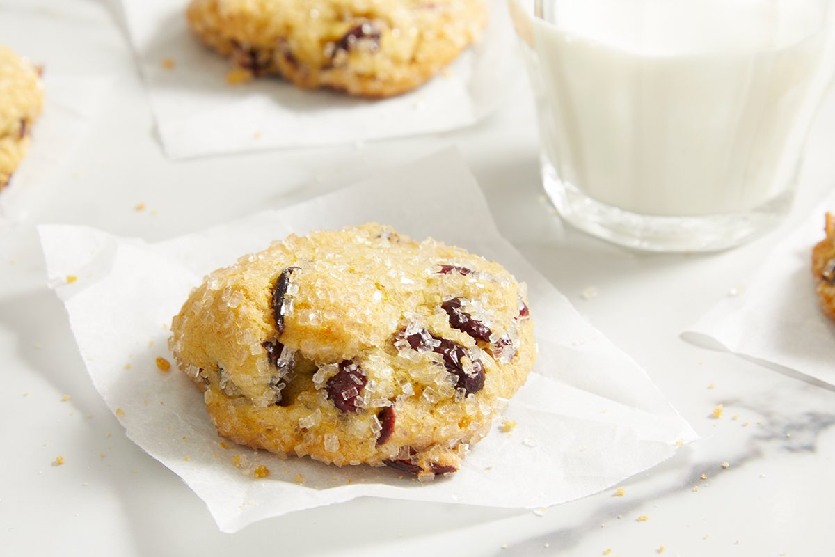 A close-up of a chocolate chip cookie on a piece of parchment paper, with a glass of milk in the background on a white marble surface.