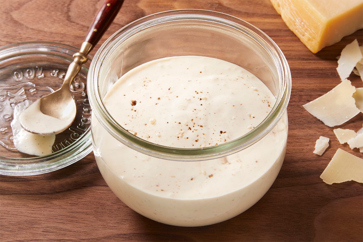 Close-up shot of Creamy Caesar dressing in a glass jar, sprinkled with cracked black pepper and surrounded by shaved Parmesan cheese.