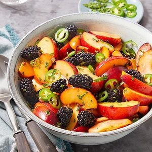 Eye Level vertical image of a bowl of peach and blackberry salad with jalapeños. The salad is garnished with fresh mint and served with salad servers.