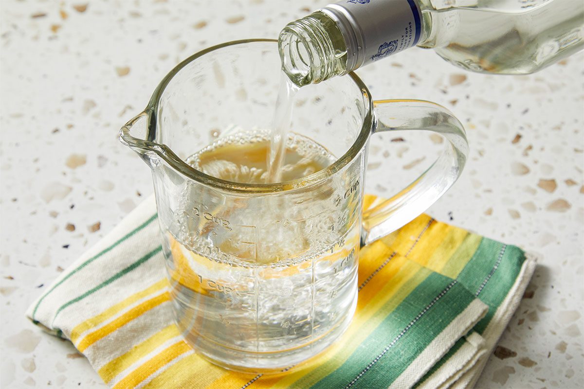 Eye level horizontal image of a clear liquid being poured from a glass bottle into a glass measuring cup. The liquid has bubbles inside the measuring cup and the measuring cup is sitting on a colorful dish towel.