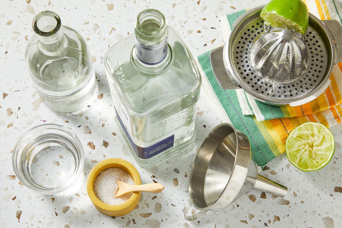 Overhead horizontal image of ingredients being prepared for a cocktail. A bottle of liquor sits next to a bowl of salt and a glass of water. A lime is being juiced on a squeezer.