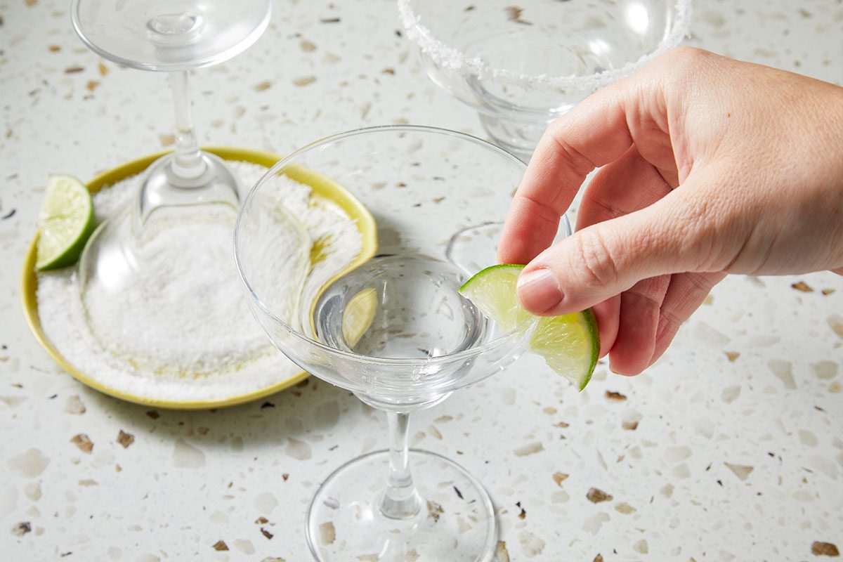 Eye Level horizontal image of a hand placing a lime wedge on a martini glass filled with clear liquid. Another martini glass with salt on the rim is visible along with a plate of salt with an upside-down glass and a lime wedge.