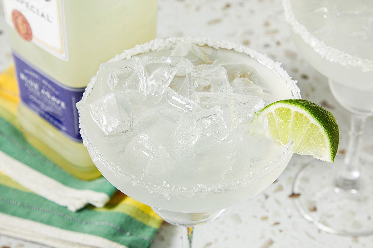 Overhead horizontal image of a margarita, rimmed with salt and garnished with a lime wedge. The drink is filled with ice cubes and a bottle of liquor is in the background.