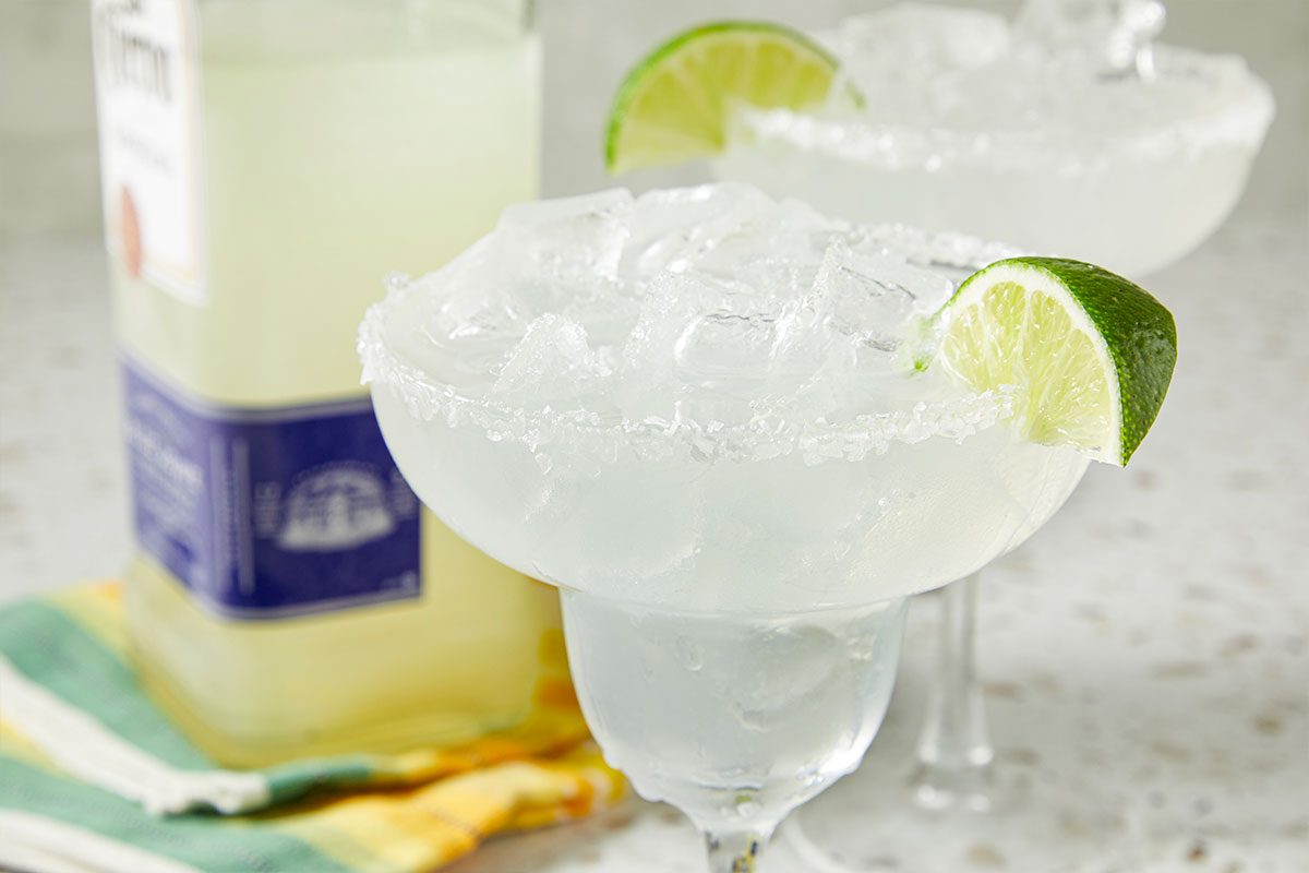 Eye Level horizontal image of margaritas being made. The rims of the glasses are salted, and the glasses are filled with ice and a lime wedge for garnish. The bottle for the drink is visible behind the glasses.