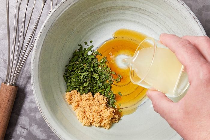 Overhead horizontal image of ingredients being added to a ceramic bowl. A hand pours liquid from a glass into the bowl which already contains chopped mint and brown sugar.
