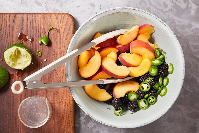 Overhead horizontal image of metal tongs moving peach slices into a bowl with blackberries and sliced jalapenos. There is a cut lime on a wooden board next to the bowl.