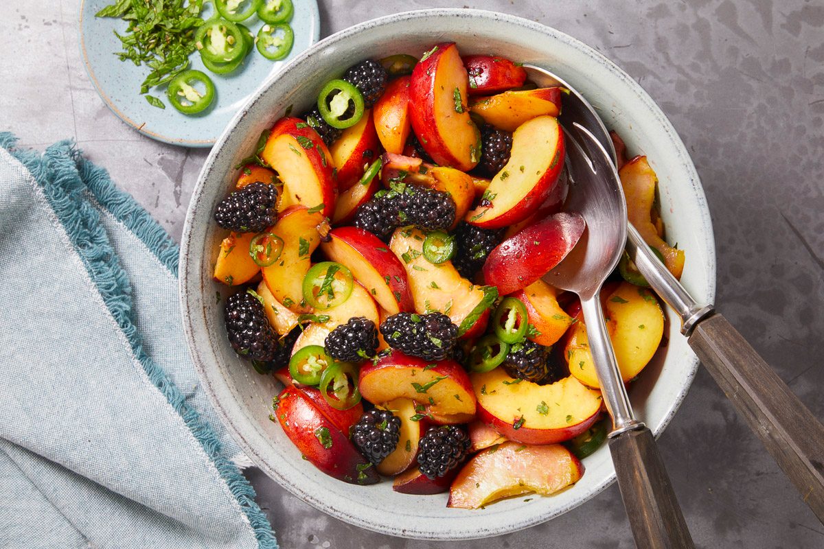 Overhead horizontal image of a bowl of peach and blackberry salad being tossed with serving spoons. Sliced jalapeños and chopped mint are mixed throughout the salad.
