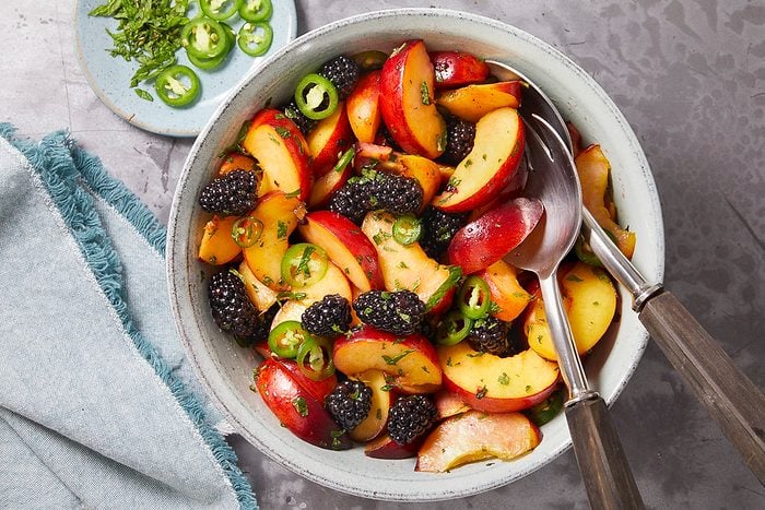 Overhead horizontal image of a bowl of peach and blackberry salad being tossed with serving spoons. Sliced jalapeños and chopped mint are mixed throughout the salad.
