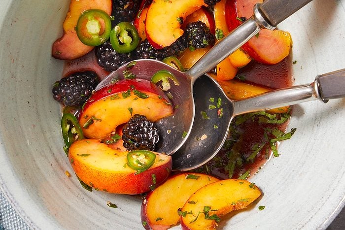 Overhead horizontal image of peach and blackberry salad with sliced jalapeños, garnished with basil and tossed with a reddish sauce. Two spoons appear in the bowl with the ingredients.