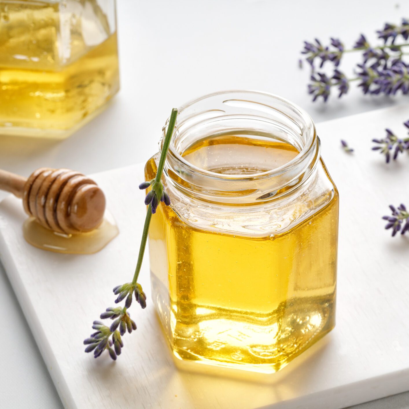 Lavender honey in opened glass jar with wooden honey dipper, drizzler and lavender flowers on a white board and background.
