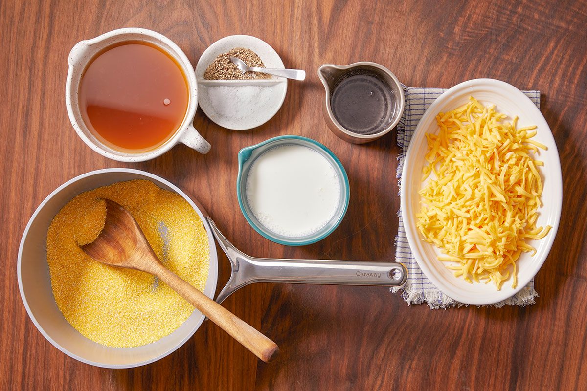 Overhead horizontal image of ingredients for polenta. The ingredients include polenta in a Caraway pan, beef broth, salt and pepper, water, milk and shredded cheese.