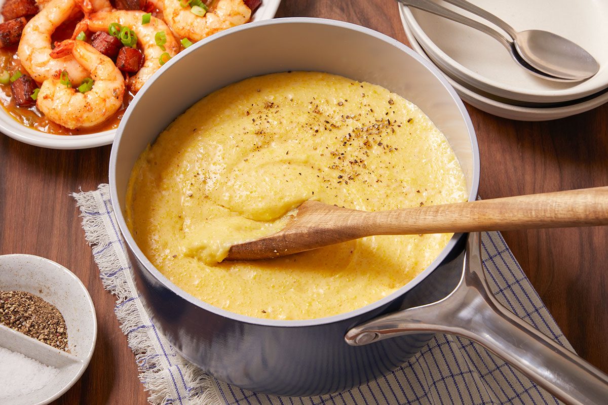 Eye Level horizontal image of creamy polenta in a blue pot with a wooden spoon resting inside. A plate of shrimp and andouille sausage is in the background, with salt and pepper in a small bowl.