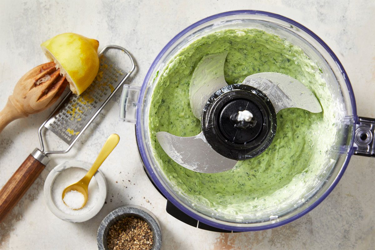 Overhead horizontal image of ingredients being processed in a food processor. Lemon, salt, and pepper are staged next to the food processor.
