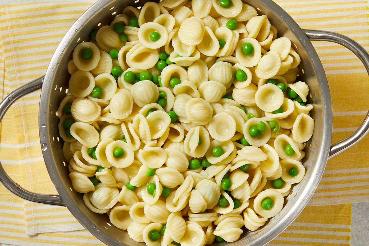 Overhead horizontal image of orecchiette pasta with peas inside a metal colander. The colander sits on a yellow and white striped towel.