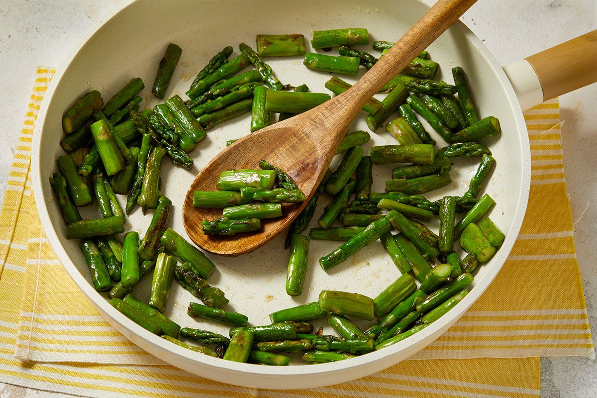 Overhead horizontal image of asparagus being cooked in a pan with a wooden spoon. The chopped asparagus pieces are coated in oil and are turning a slightly darker shade of green as they cook.
