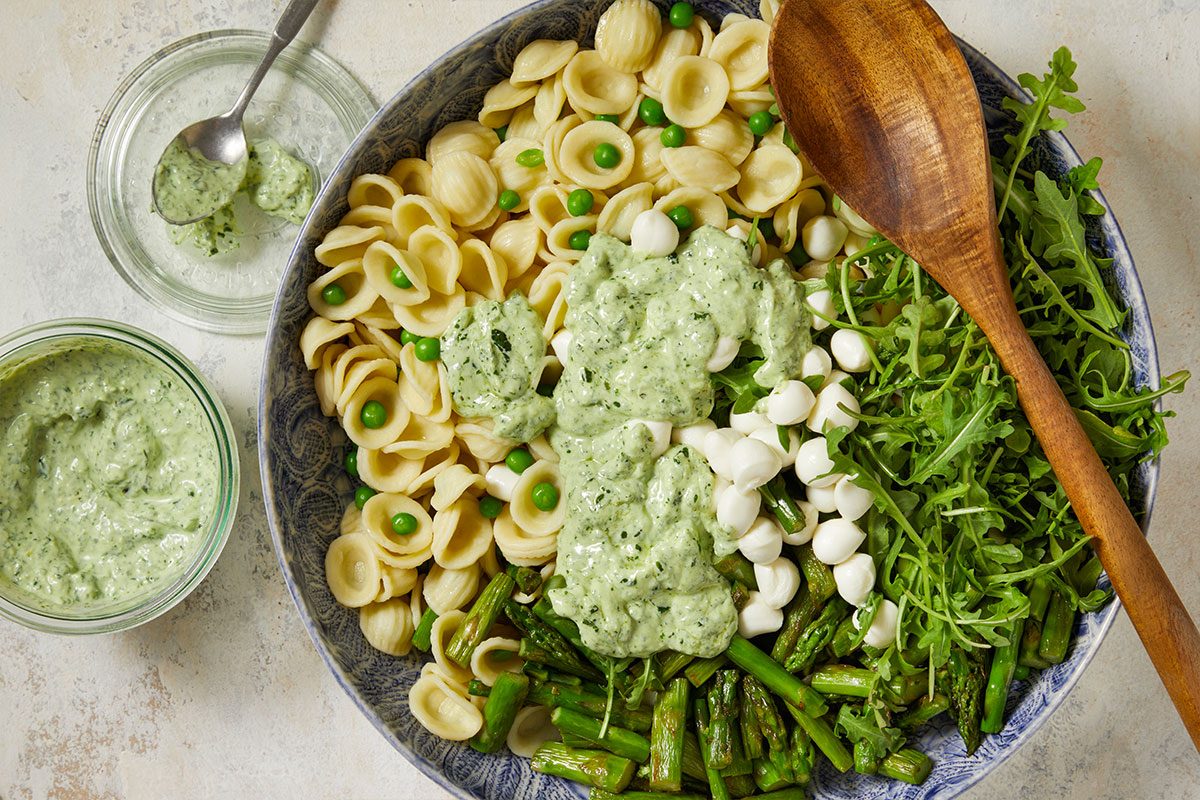 Overhead horizontal image of a large blue patterned bowl filled with orecchiette pasta, peas, mozzarella balls, arugula, and cooked asparagus. The salad is topped with green pesto sauce, and the bowl sits on a white surface next to a jar of pesto sauce and a wooden serving spoon.