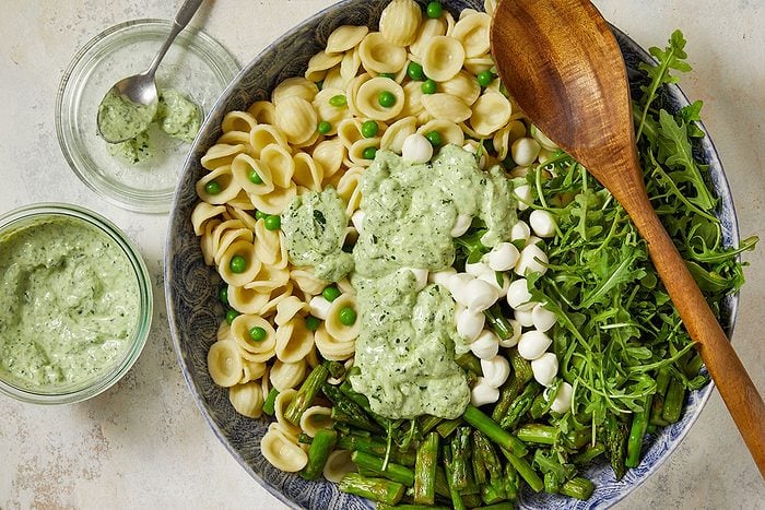 Overhead horizontal image of a large blue patterned bowl filled with orecchiette pasta, peas, mozzarella balls, arugula, and cooked asparagus. The salad is topped with green pesto sauce, and the bowl sits on a white surface next to a jar of pesto sauce and a wooden serving spoon.