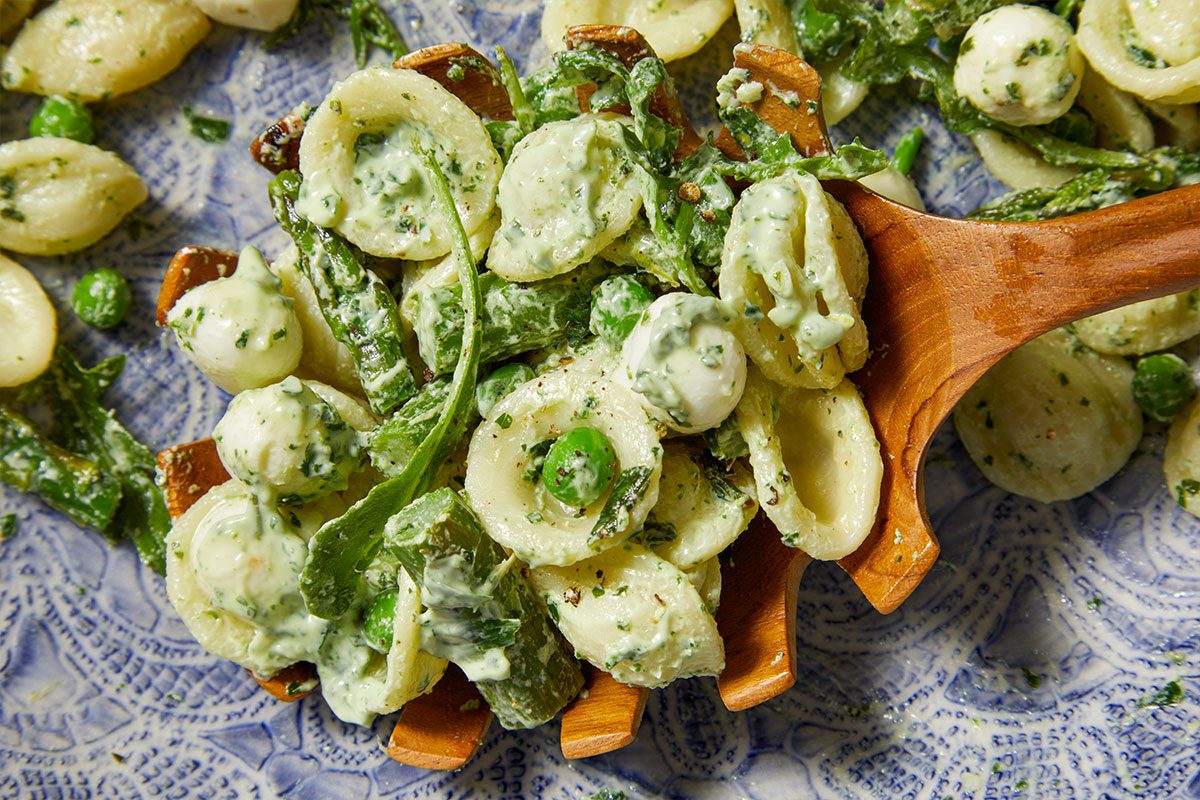 Overhead horizontal image of Orecchiette pasta tossed with a creamy, pesto-like sauce on a wooden serving spoon. The dish also includes bocconcini mozzarella, green peas, and asparagus.