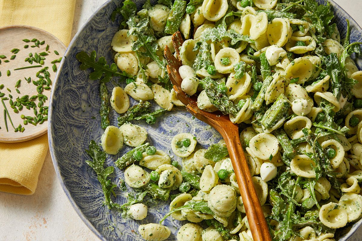 Overhead horizontal image of orecchiette pasta salad in a blue patterned bowl with a wooden serving spoon. The pasta salad includes peas, asparagus, arugula and small mozzarella balls. Chopped chives are visible on a small wooden plate nearby.