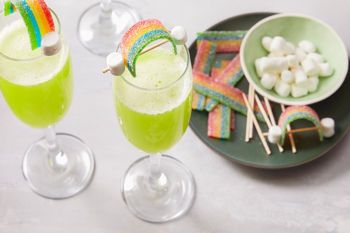 Two glasses of green drink are garnished with rainbow candy and marshmallows. Next to them is a plate with more rainbow candies, marshmallows, and toothpicks. The scene is bright and festive.