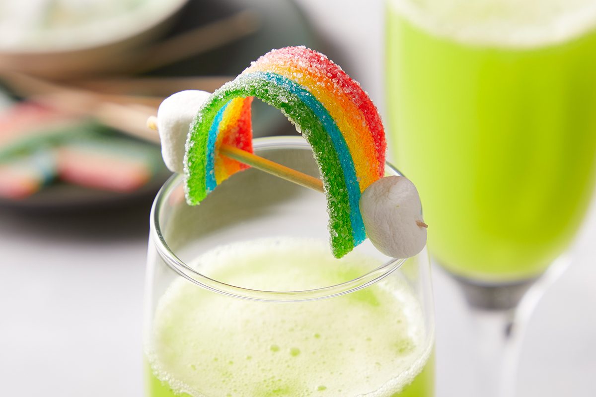 A glass of bright green drink is garnished with a candy rainbow held by two toothpicks, with mini marshmallows on each end of the rainbow. Another glass and a plate are blurred in the background.