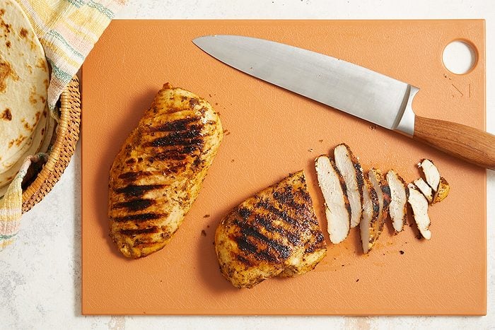 Overhead horizontal image of grilled chicken being sliced on a cutting board with a knife. To the side, in a basket, there are tortillas ready to be filled with chicken.