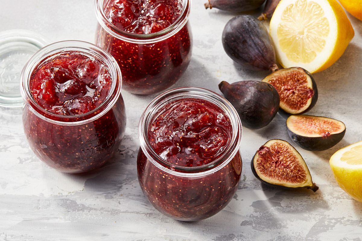 Overhead horizontal image of three jars of fig jam on a light grey surface with whole and halved figs and lemon. One jar is open and the other two are sealed. | [Original] RMS 281249 Fig Jam Recipe; Horizontal; AP Pull