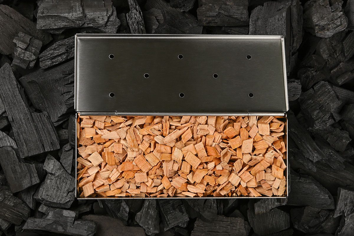 Close up one stainless steel metal smoker box with hardwood alder chips on black lump charcoal pieces, ready to barbecue grill and smoke food