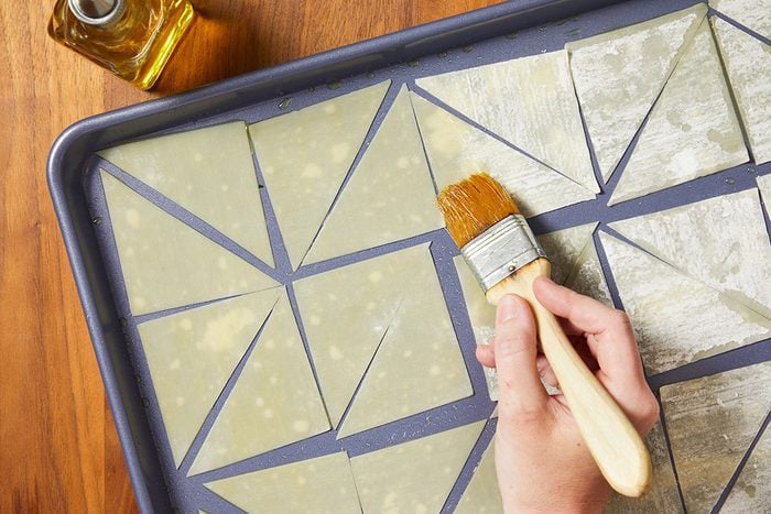 Overhead horizontal image of wonton wrappers cut into triangles on a baking sheet. A hand is shown using a pastry brush to brush the wrappers with oil.