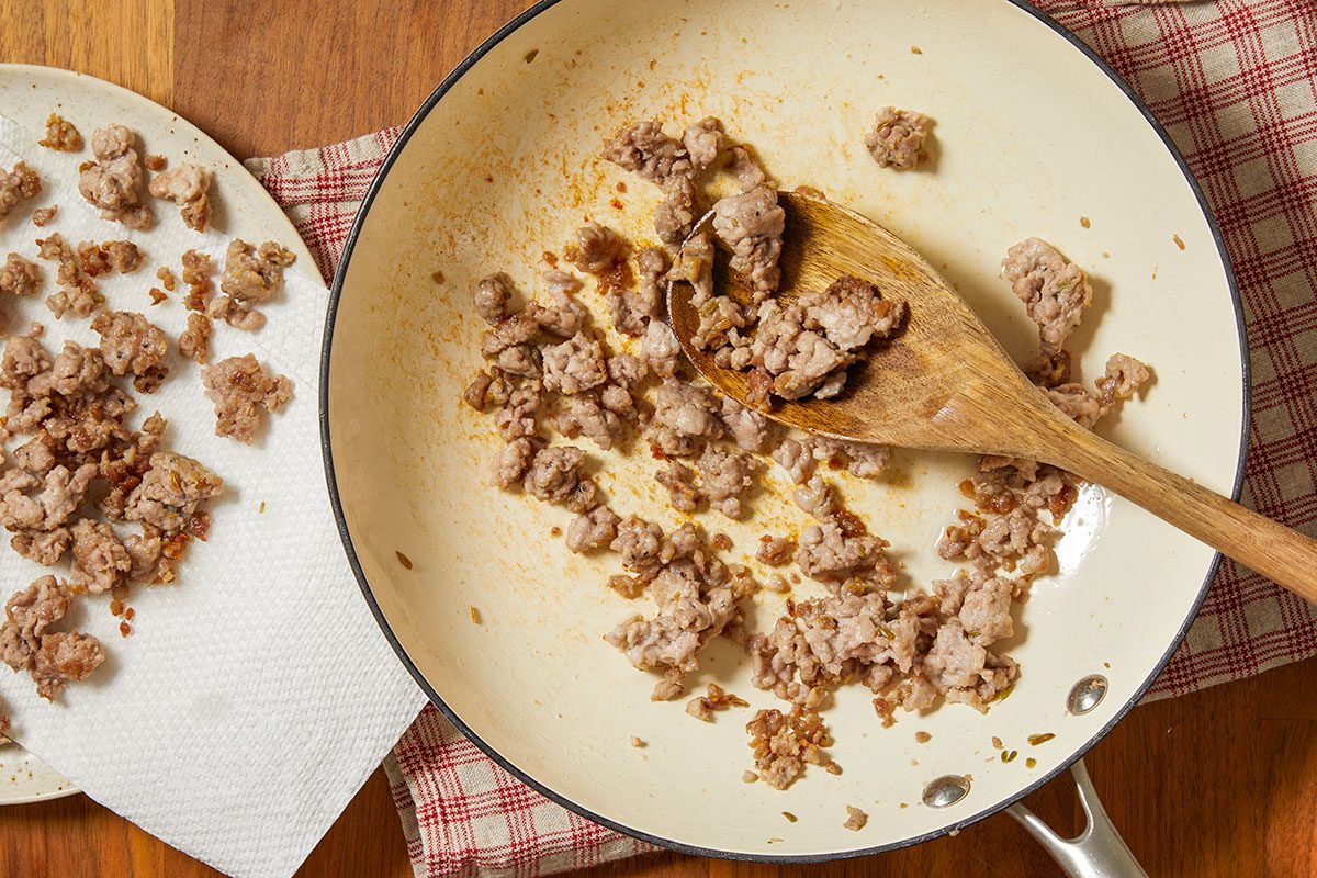 Overhead horizontal image of browned ground meat in a skillet with a wooden spoon. Some of the cooked meat is plated on a paper towel to drain off excess fat.
