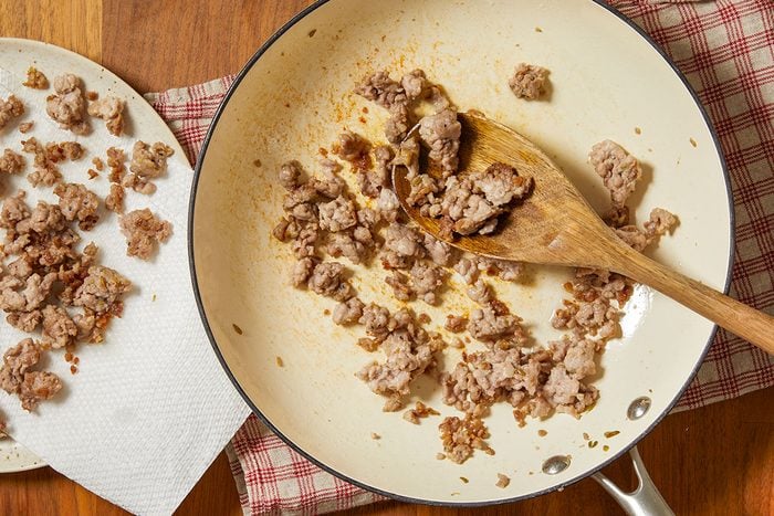 Overhead horizontal image of browned ground meat in a skillet with a wooden spoon. Some of the cooked meat is plated on a paper towel to drain off excess fat.