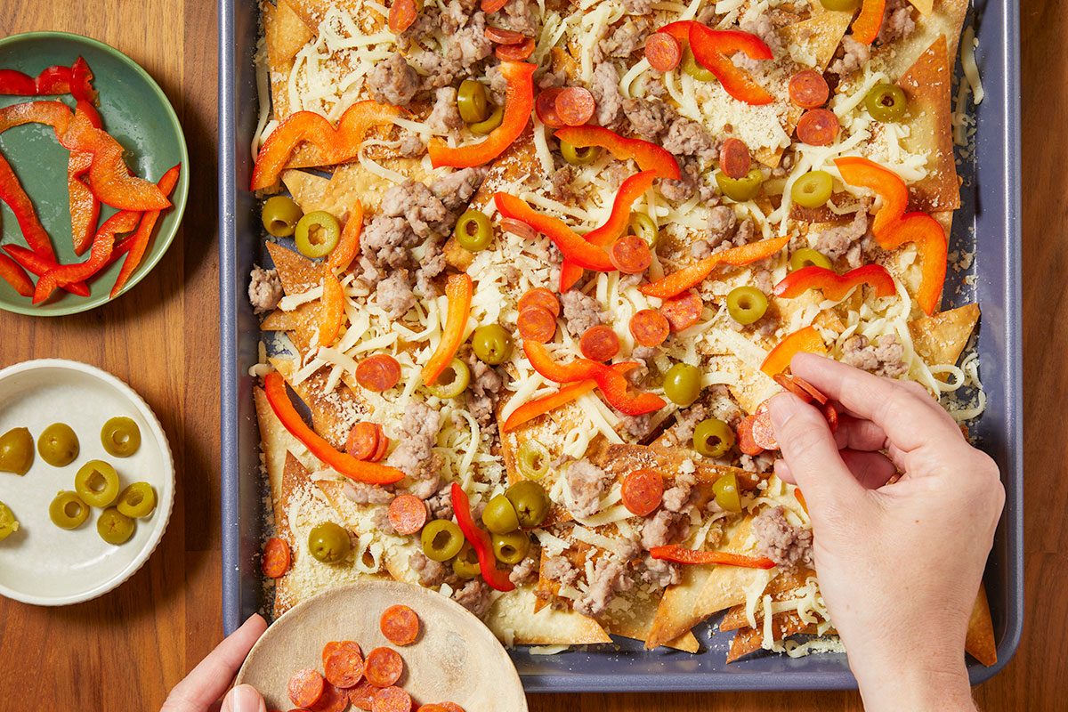 Overhead horizontal image of nachos being assembled in a baking pan. Sliced pepperoni, red bell peppers, olives, cooked ground meat and shredded cheese are being layered on top of the tortilla chips.